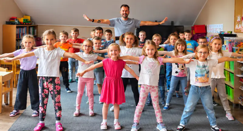 Duża grupa przedszkolaków stoi w sali z rozłożonymi rękami i powtarza ruch za instruktorem